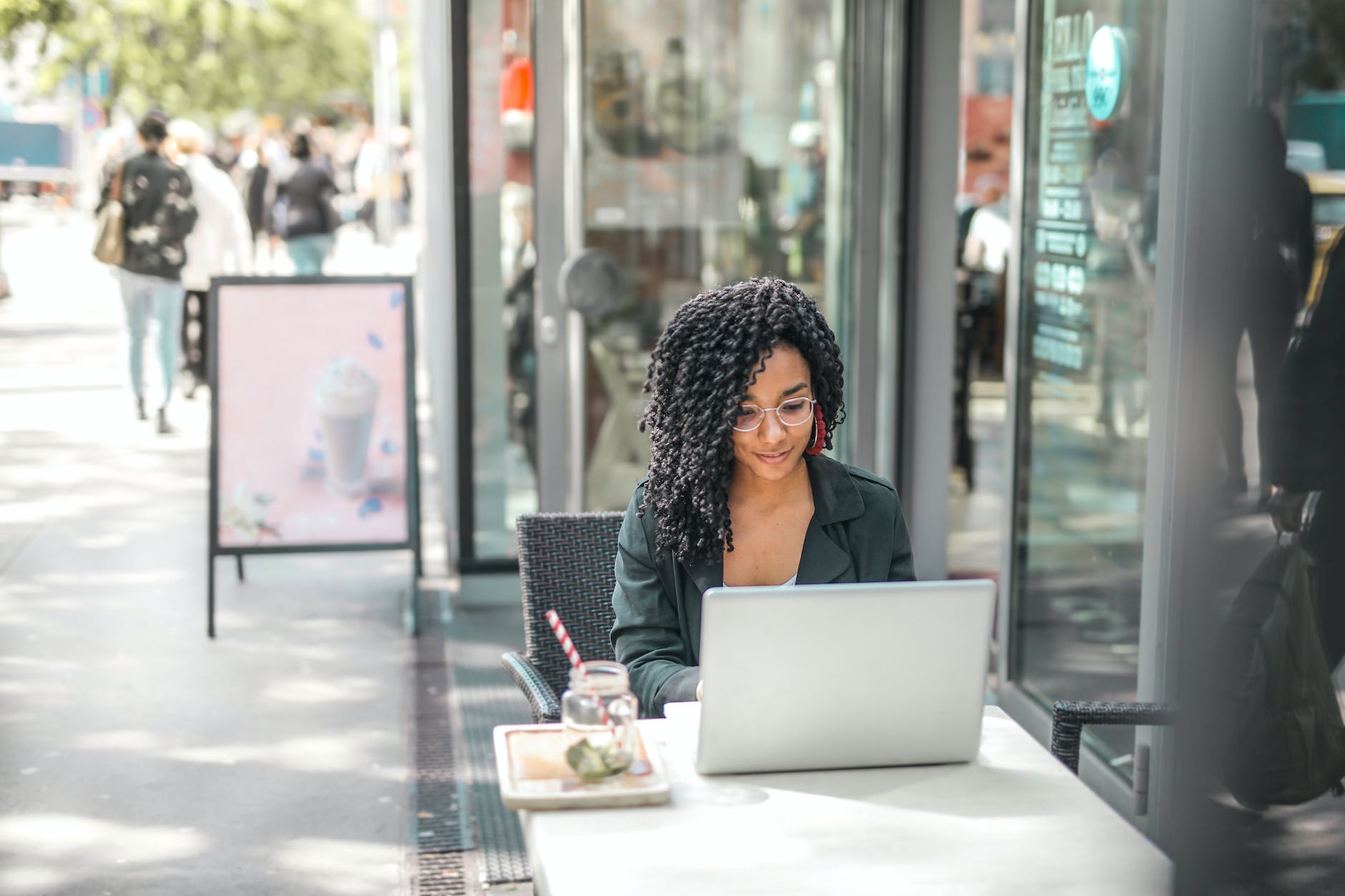 High angle of pensive African American female freelancer in glasses and casual clothes focusing on screen and interacting with netbook while sitting at table with glass of yummy drink on cafe terrace in sunny day (Photo by Andrea Piacquadio)